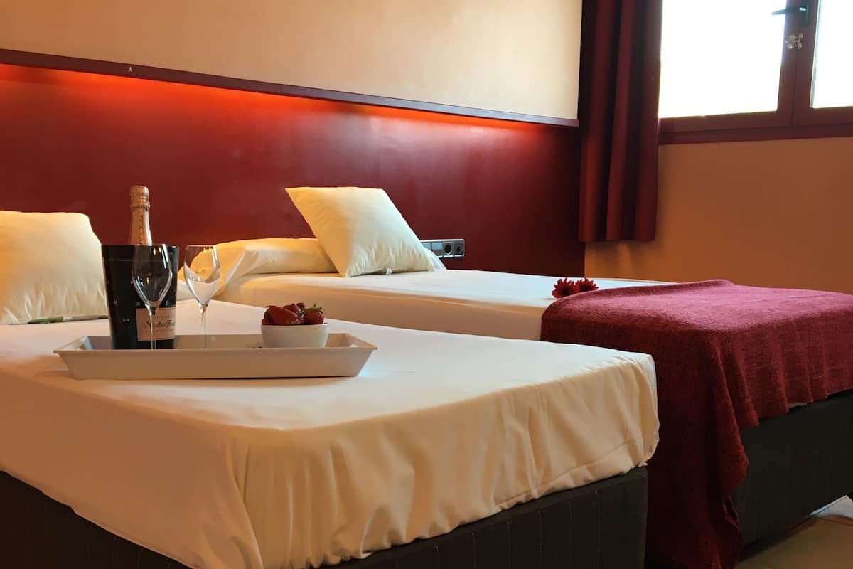 Habitación de hotel con dos camas, un atril con una botella y vasos, y un par de zapatos en el suelo.