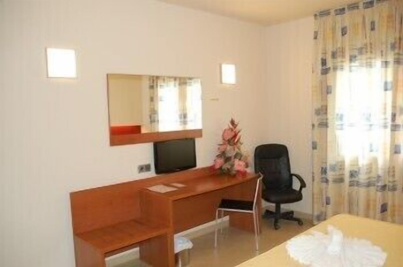 A hotel room with a wooden desk, a black office chair, a flat-screen TV, a small mirror, and a window with curtains.
