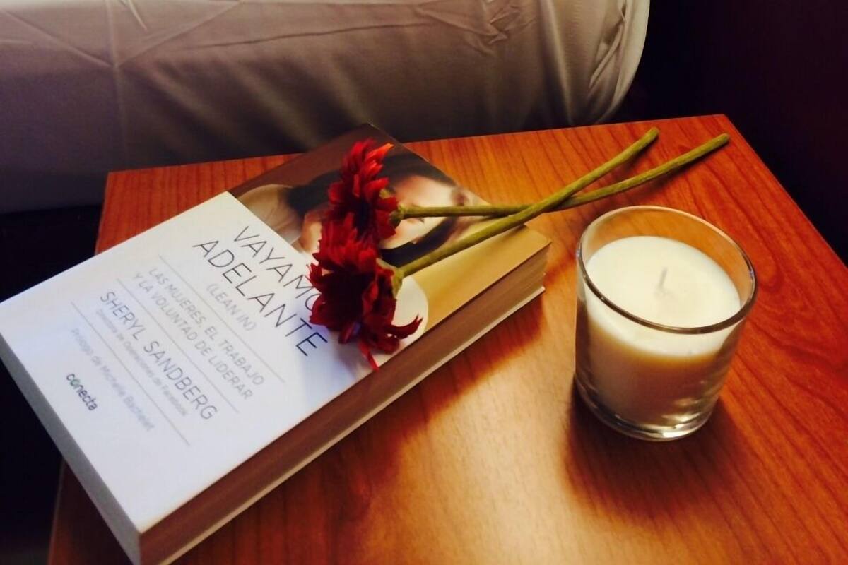 A neatly made bed with white linens, a book titled 'Vaya a la mente' by Sheryl Sandberg, a red flower, and a lit candle on a wooden side table.