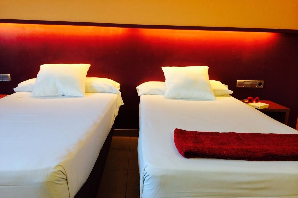 Two single beds with white linens and pillows, a red towel on the right bed, and a wall-mounted control panel.