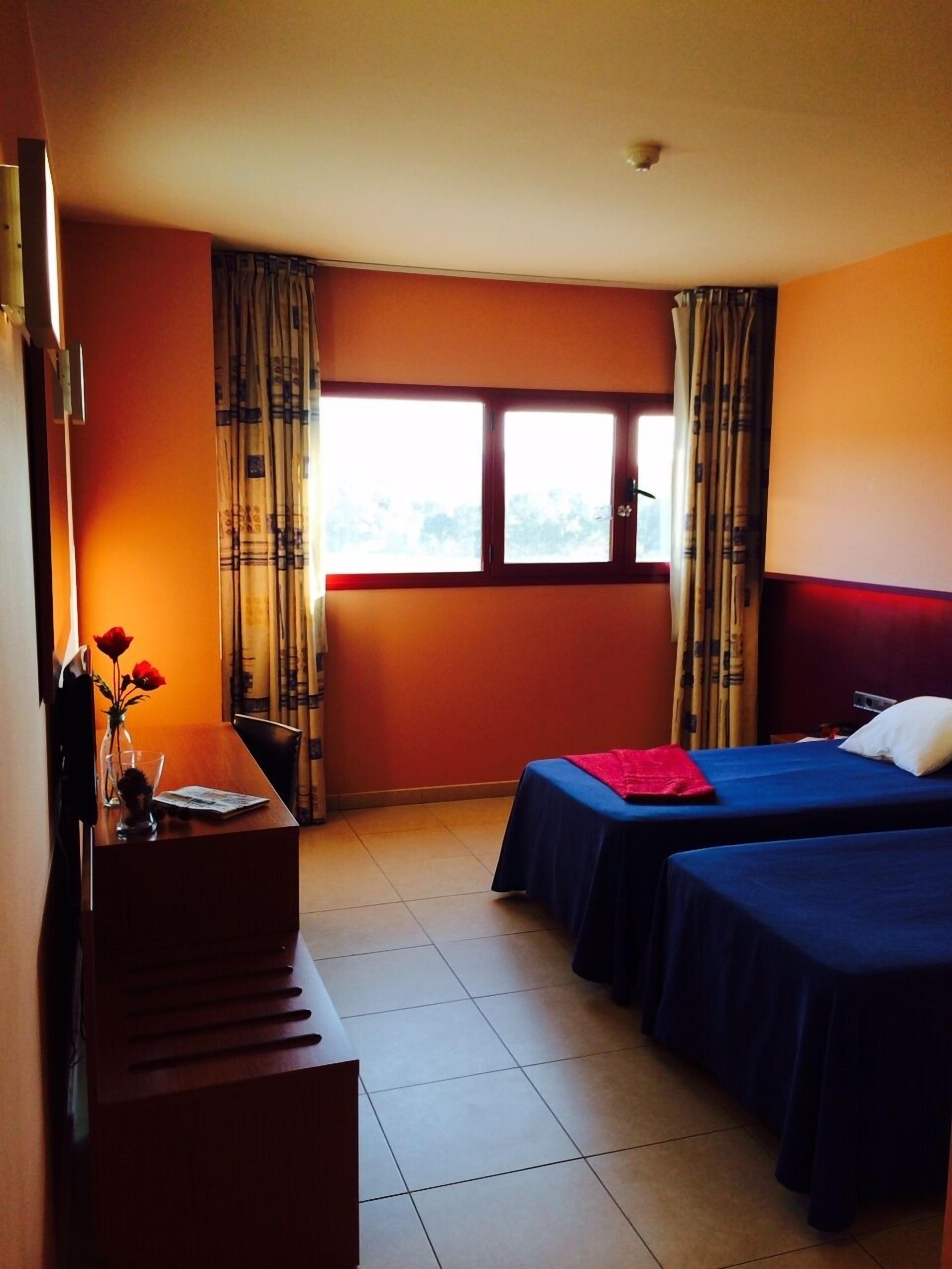 Habitación de hotel con dos camas, un escritorio, una silla, un ventanal con cortinas y un florero con flores.