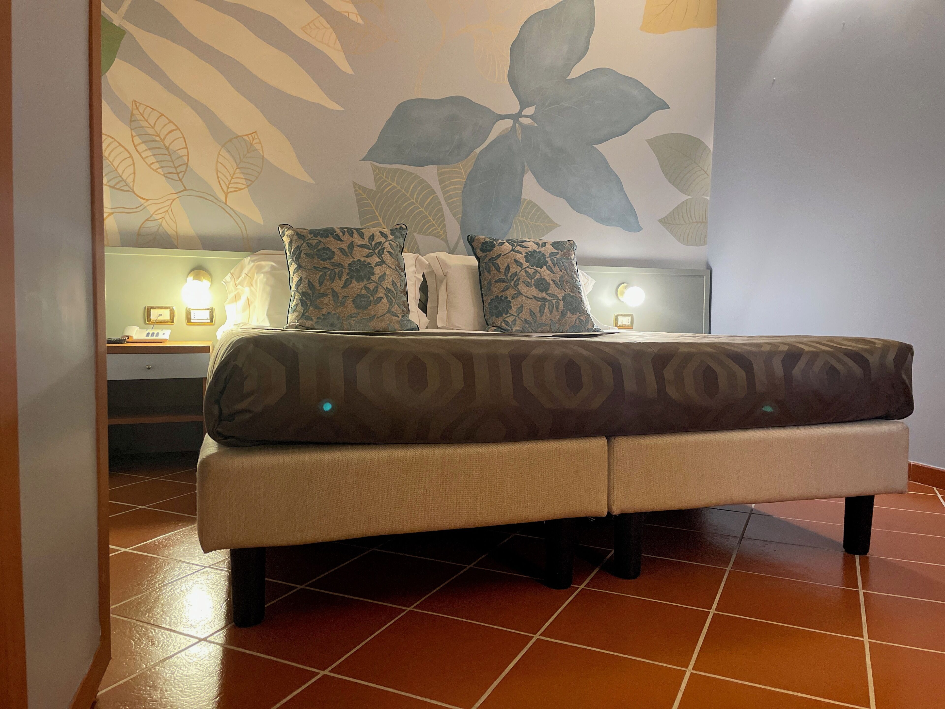 A bedroom with a bed, two pillows, a bedside table with a lamp, and a wall with a leaf pattern.