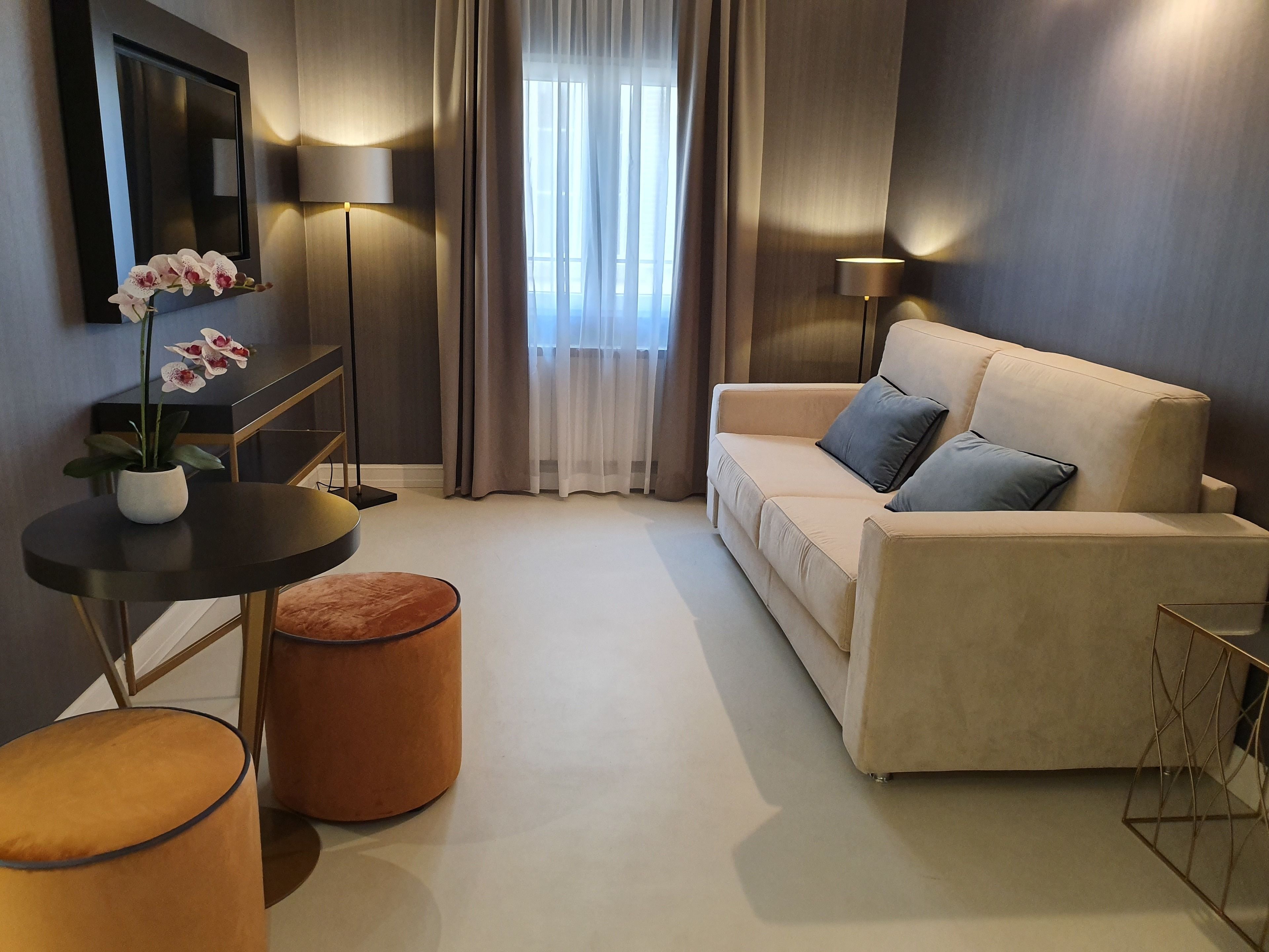 A modern living room with a beige sofa, two side tables, a small orange ottoman, a potted orchid, and a large window with sheer curtains.
