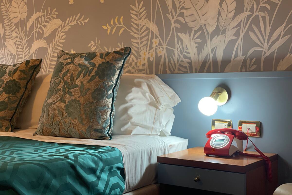 Hotelkamer met een bed, kussens, een nachtkastje met een rode draaischijf telefoon en een muur met bloemen behang.