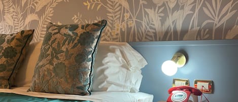 Habitación de hotel con cama, almohadas, una mesita de noche con teléfono giratorio rojo y un papel tapiz floral en la pared.