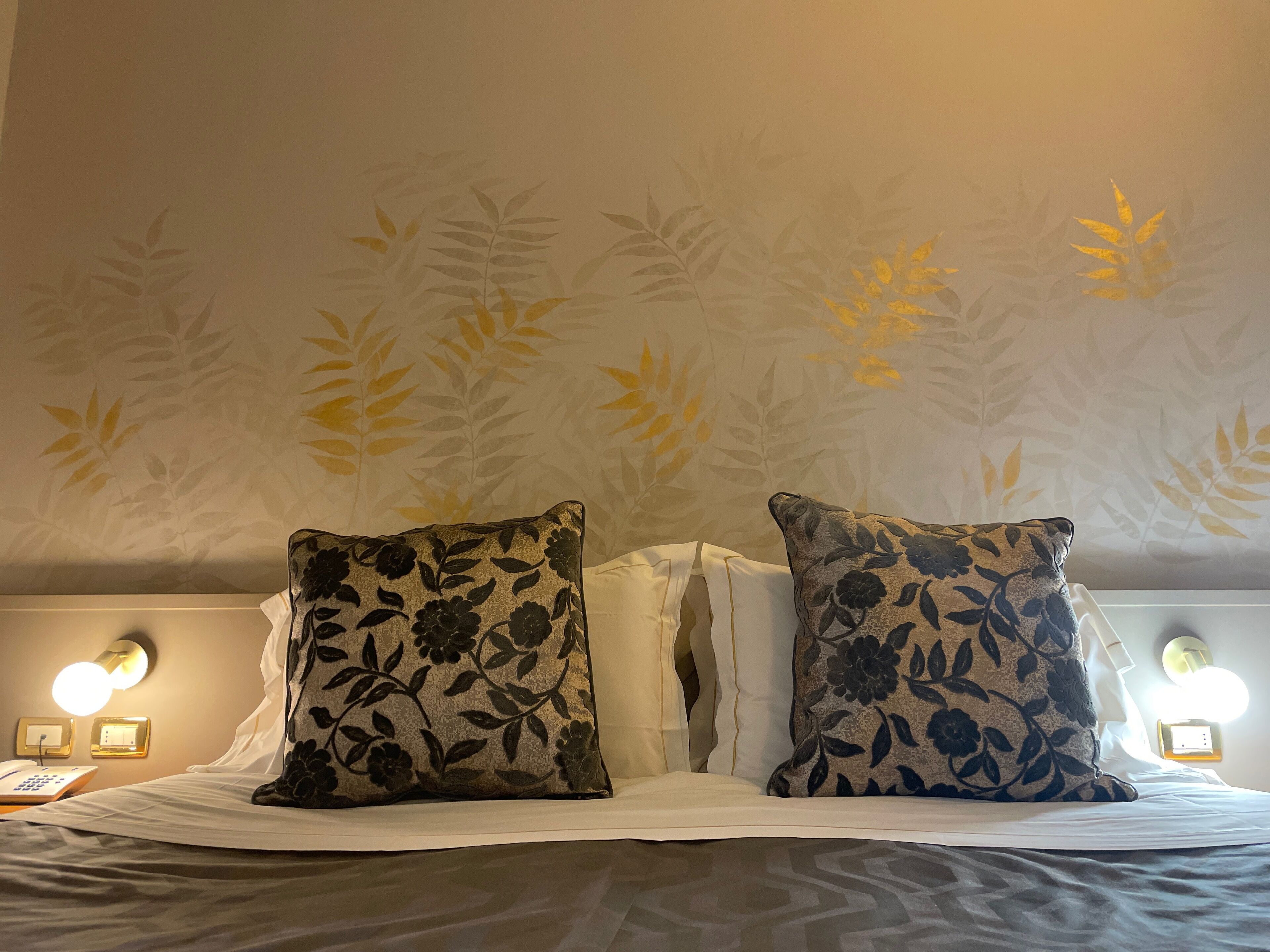 Una cama con dos almohadas de estampado floral, un sábana blanca y una pared con diseño de hojas.