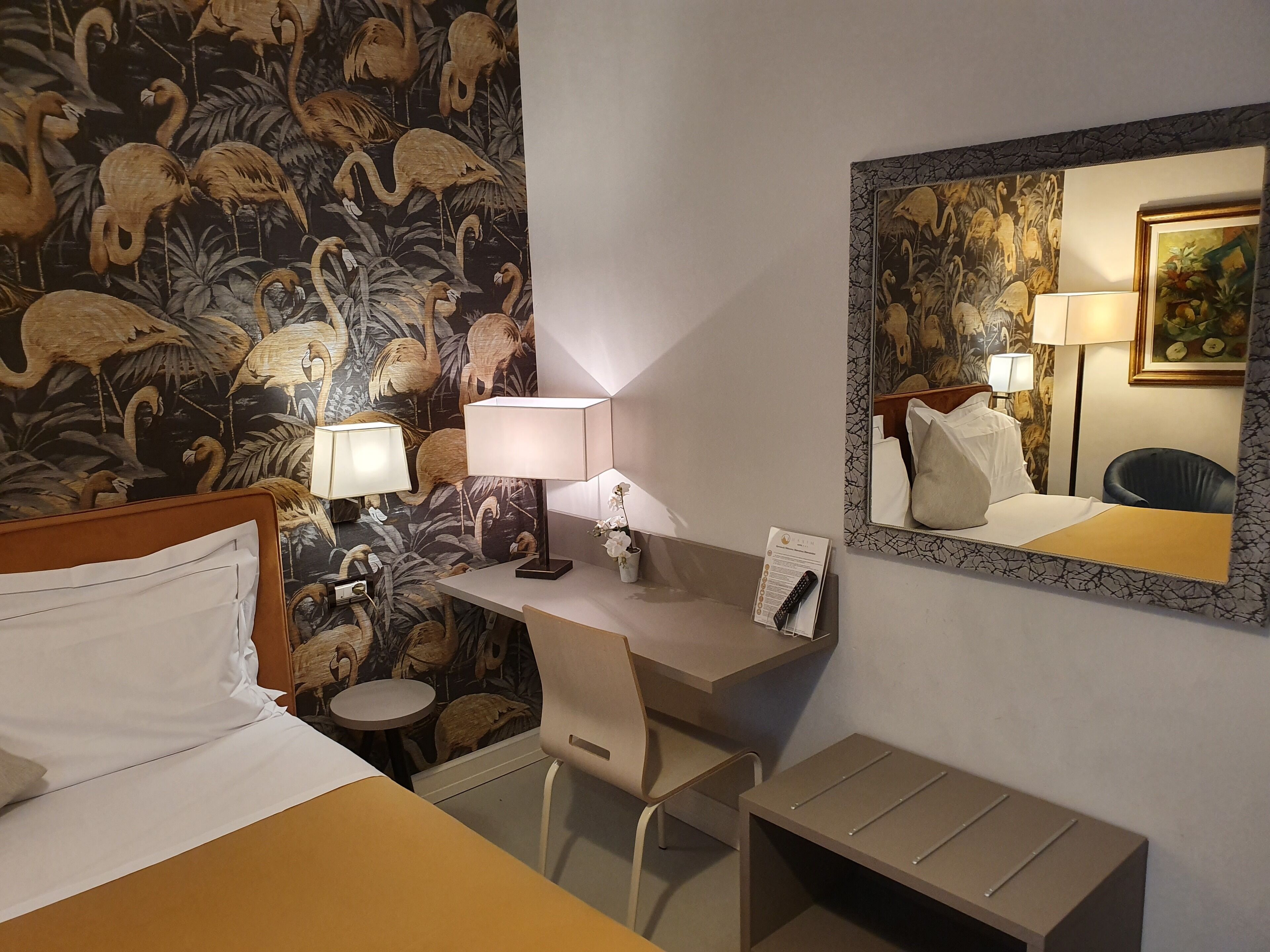 Ein Hotelzimmer mit Bett, Schreibtisch, Stuhl und einem kleinen Tisch. An der Wand hängt ein Spiegel, auf dem Schreibtisch eine Lampe und darüber ein Bild.