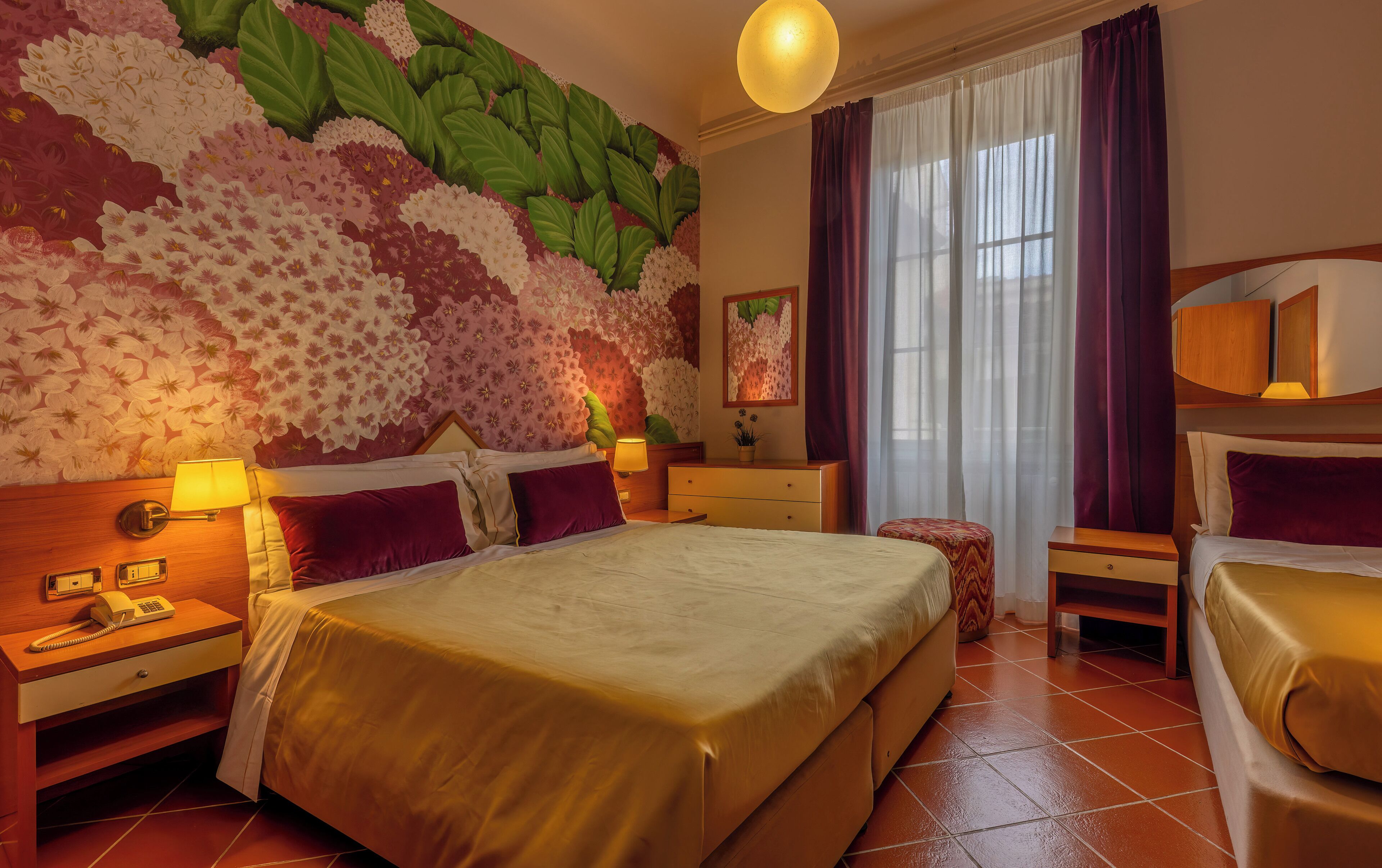 A hotel room with a large bed, a bedside table with a lamp, a nightstand, a dresser, and a large window with curtains.