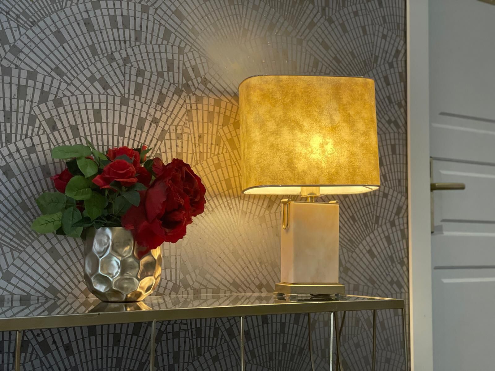 Une table avec une lampe et un vase de roses rouges, le tout placé devant un mur à motifs.
