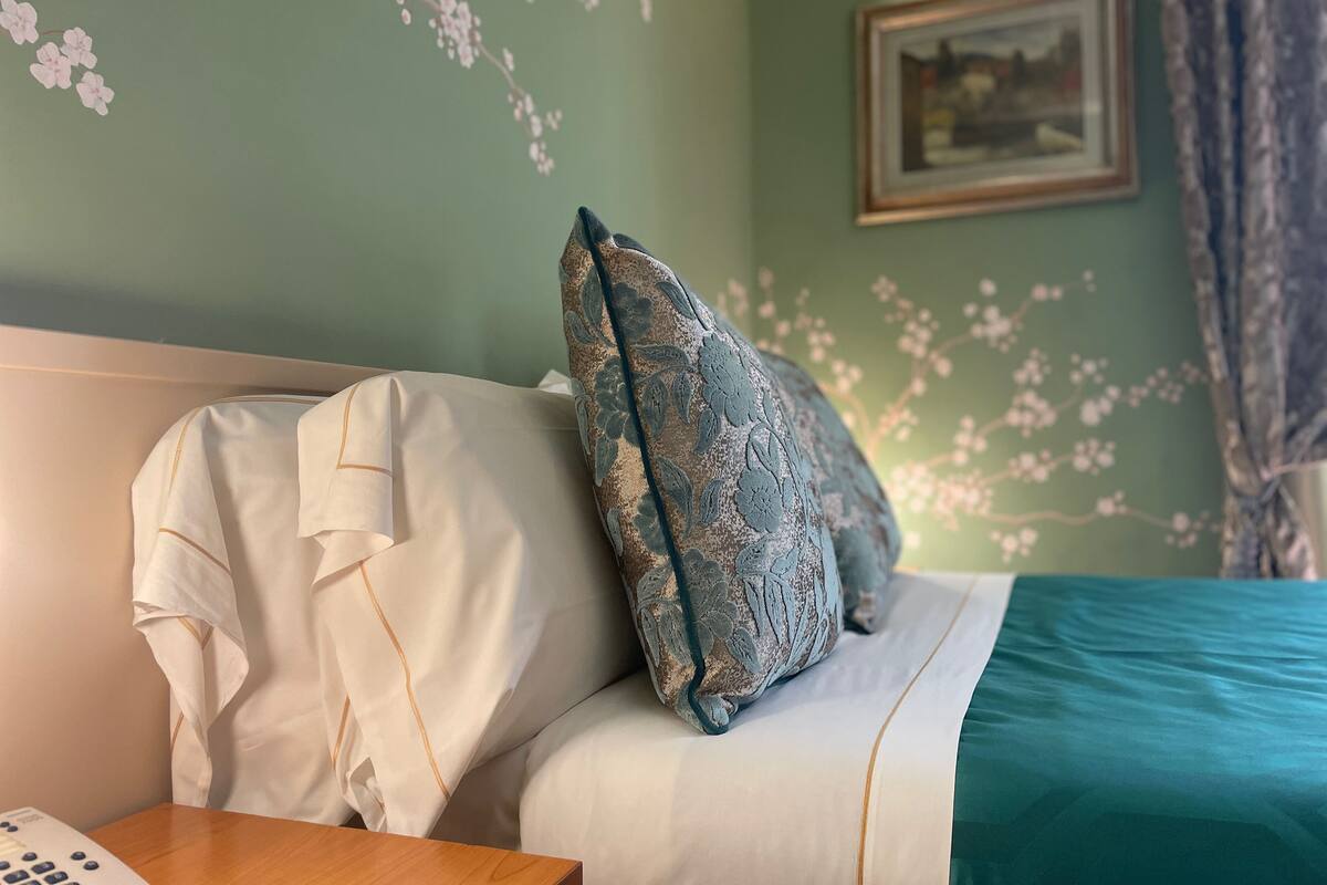 Een netjes opgemaakt bed met een blauwgroene sprei, een wit kussen met een bloemenpatroon en een wit kussen met gouden rand.