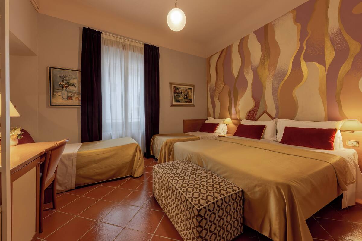 Een hotelkamer met twee bedden, een bureau en een stoel. Twee schilderijen hangen aan de muur, een raam met gordijnen en een gestreept voetenbankje.