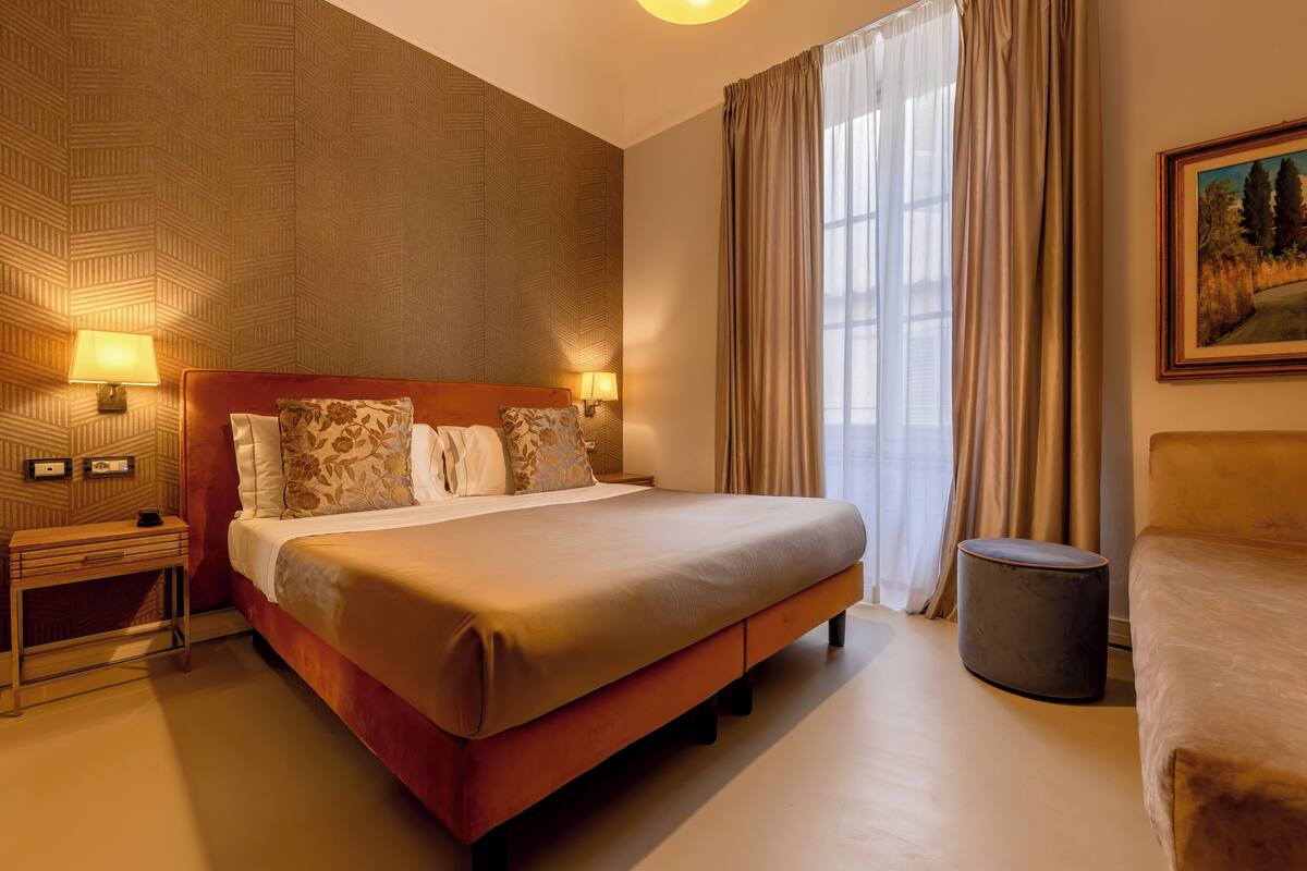 Een hotelkamer met een groot bed, twee nachtkastjes met lampen, een rond poef en een schilderij aan de muur.