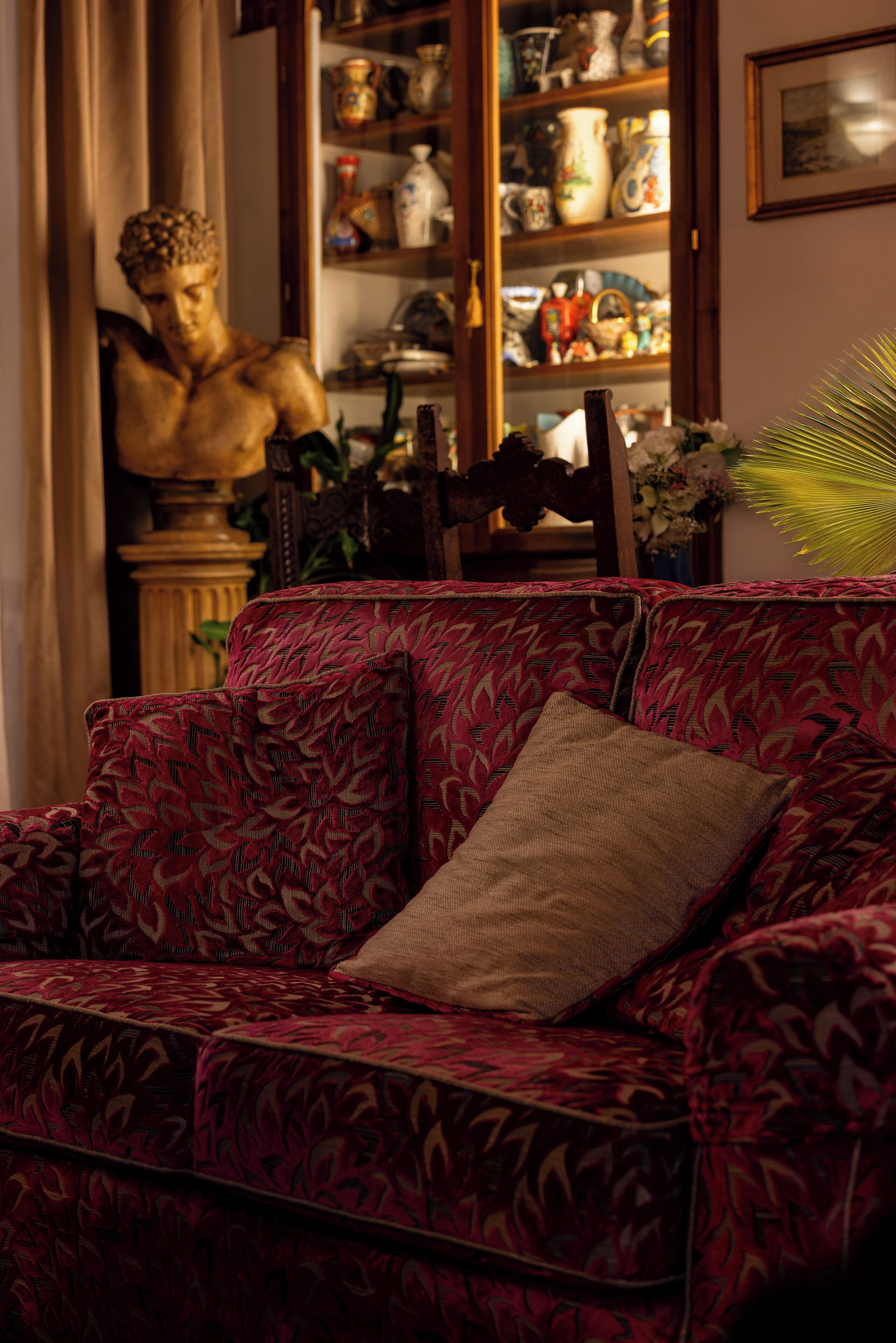 Un divano rosso con motivi decorativi, un cuscino beige, una sedia in legno e una statua sullo sfondo.