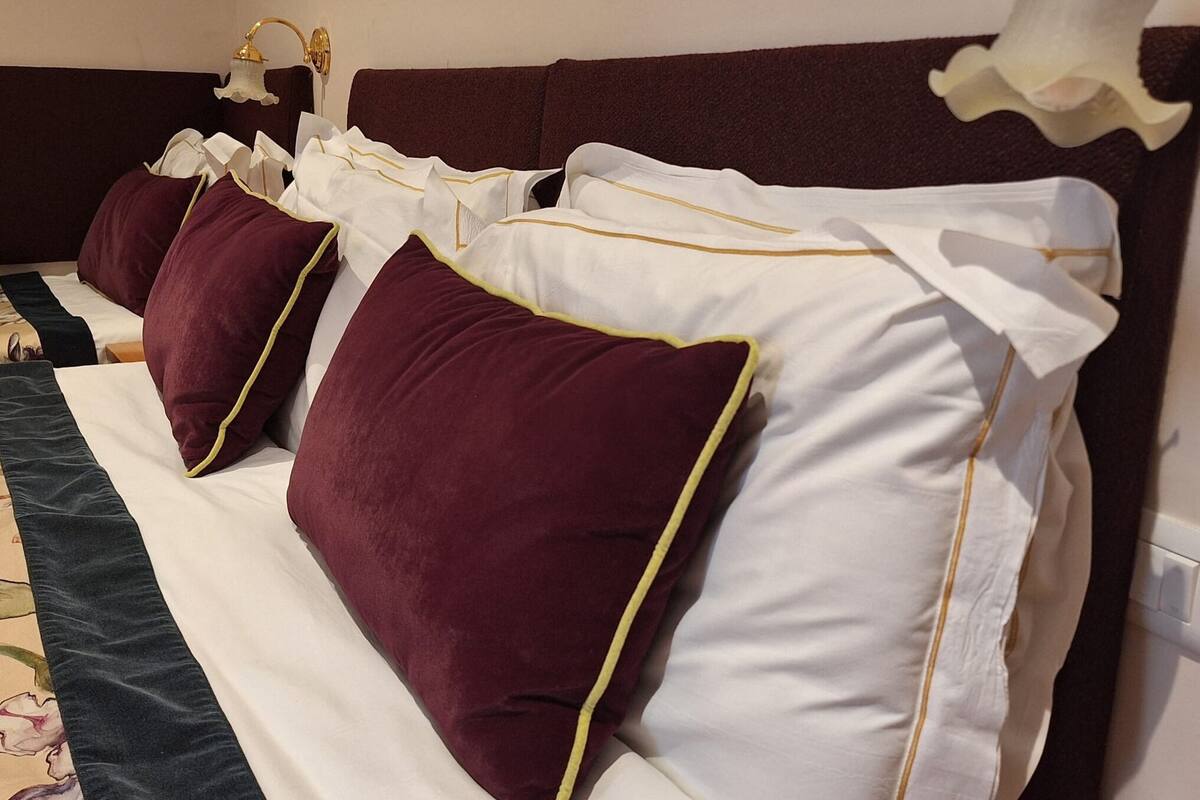 Een hotelkamer met een bed, voorzien van witte en bordeauxrode kussens, een donker hoofdeinde en een wandlamp.