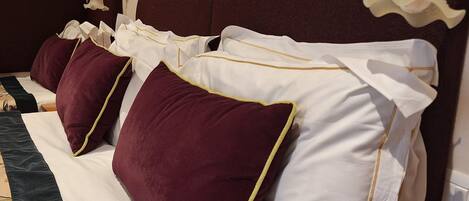 A hotel room with a bed featuring white and maroon pillows, a dark headboard, and a wall-mounted lamp.