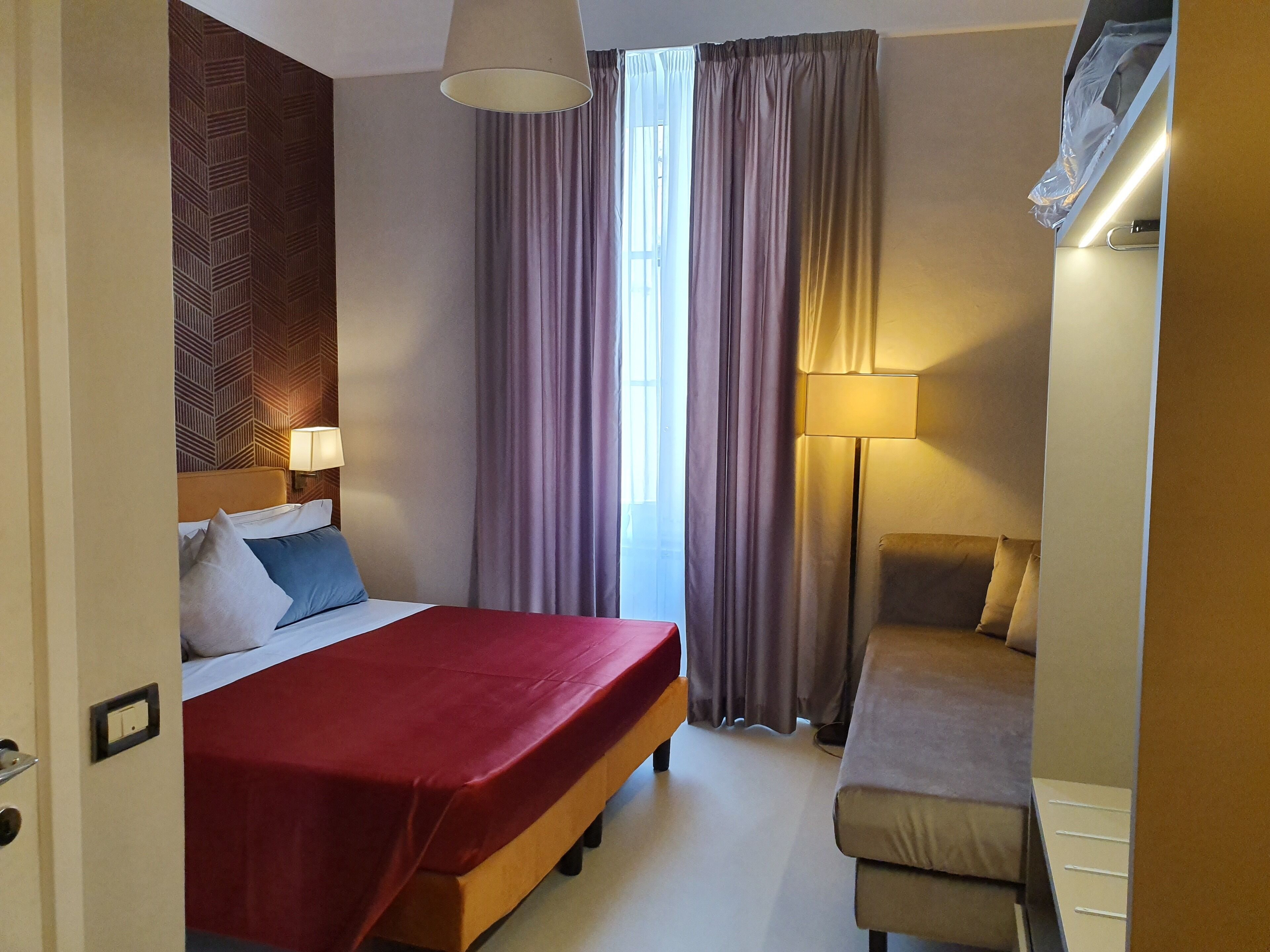 A hotel room with a bed, bedside table, lamp, curtains, and a sofa.