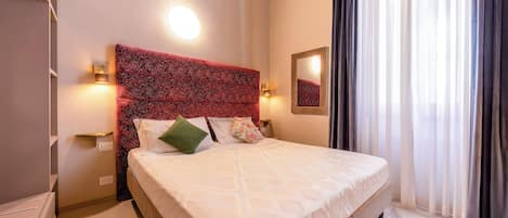 A hotel room with a large bed, a red textured headboard, two bedside lamps, a mirror, and a window with curtains.