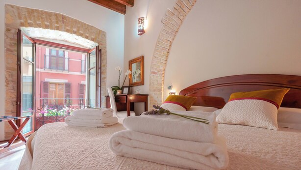 A hotel room with a large bed, a wooden headboard, and a balcony with a view of a building. There are towels on the bed and a vase with flowers.