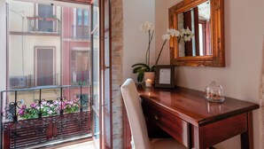 A room with a wooden desk, a mirror, a vase with flowers, and a window with a view of balconies.