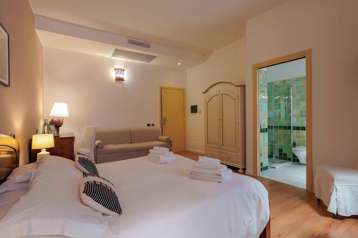 A hotel room with a large bed, a sofa, a wardrobe, and a bathroom visible through a glass door.