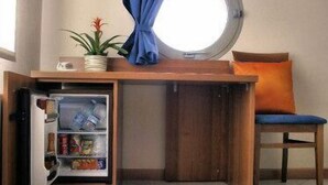 A compact room with a built-in refrigerator, a small table, a chair with an orange cushion, and a window with a curtain. - Albergo Meublè Abatjour (Mantua)