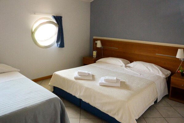 A hotel room with a single bed, a bedside table with a lamp, a round window with curtains, and a small table with a flower vase.