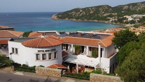 Un hotel costero de fachada blanca y tejados de tejas rojas, llamado "HOTEL MON LEPOS". Cuenta con una terraza con mobiliario exterior y vistas al mar y un paisaje montañoso al fondo.