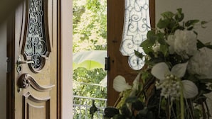 Una puerta de madera abierta con detalles de metal intrincados, que da a un balcón con una planta en maceta y vistas al verdor.
