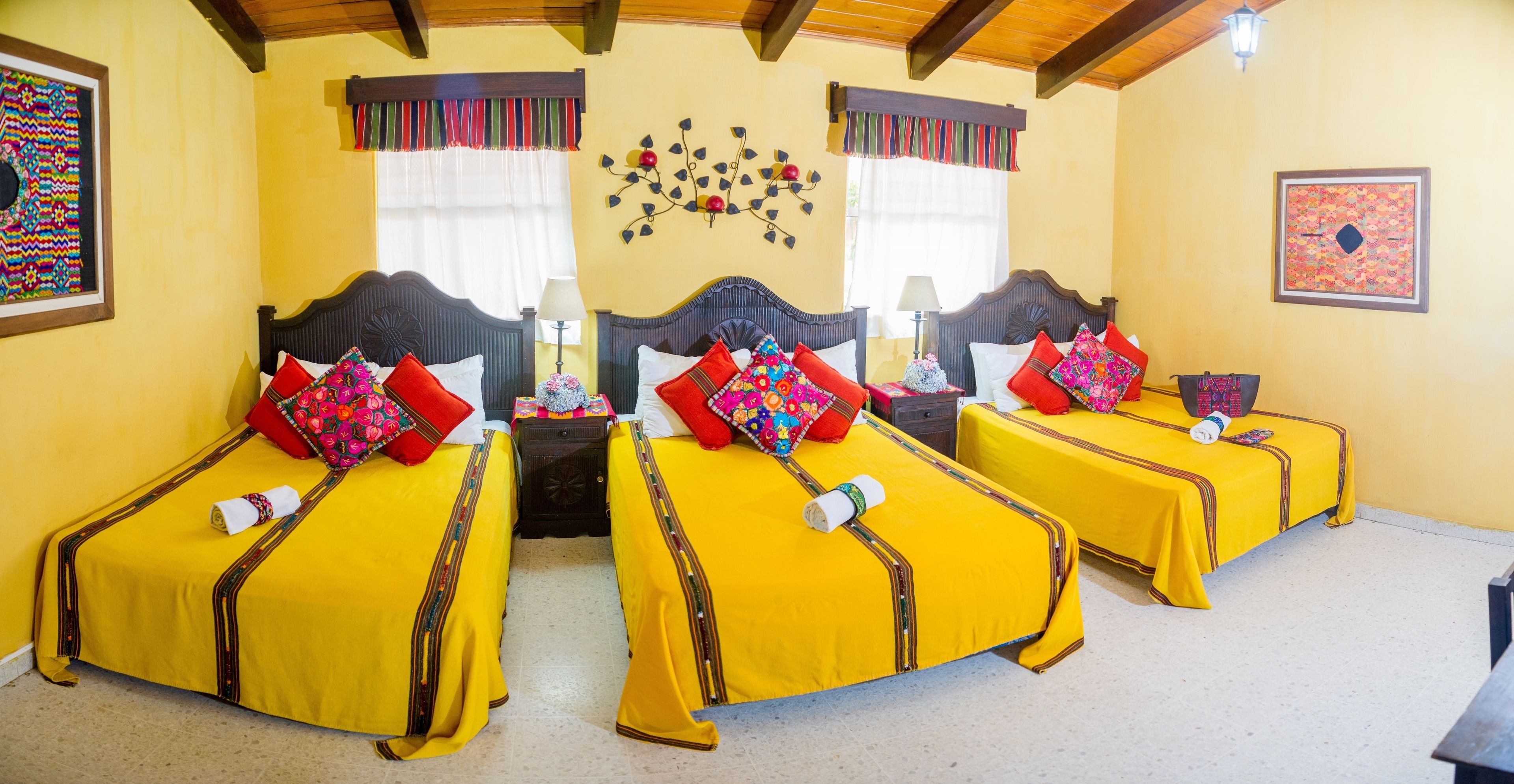 Quarto de hotel com duas camas, cada uma com roupa de cama amarela e almofadas vermelhas. Possui duas mesas de cabeceira com luminárias e objetos decorativos. O quarto tem vigas de madeira no teto e uma parede com uma obra de arte floral.