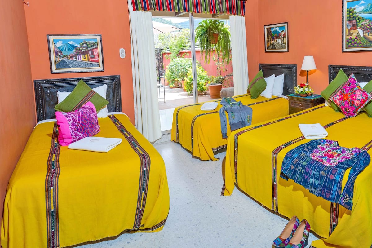Habitación de hotel con tres camas, cada una con una colcha amarilla y cojines decorativos. Se ve una ventana con vistas al exterior, una puerta que da a un balcón y un cuadro en la pared.