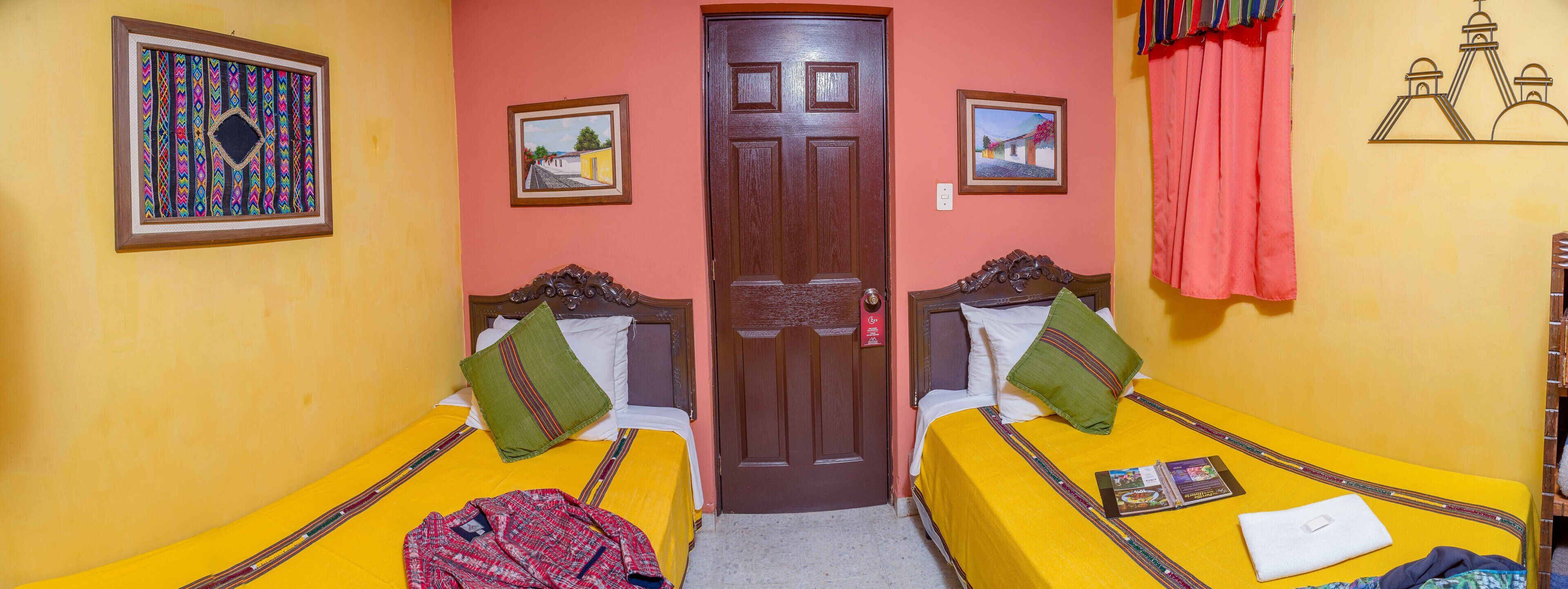 A room with two beds, each with a yellow bedspread and pillows, a wooden door, and two framed pictures on the walls.