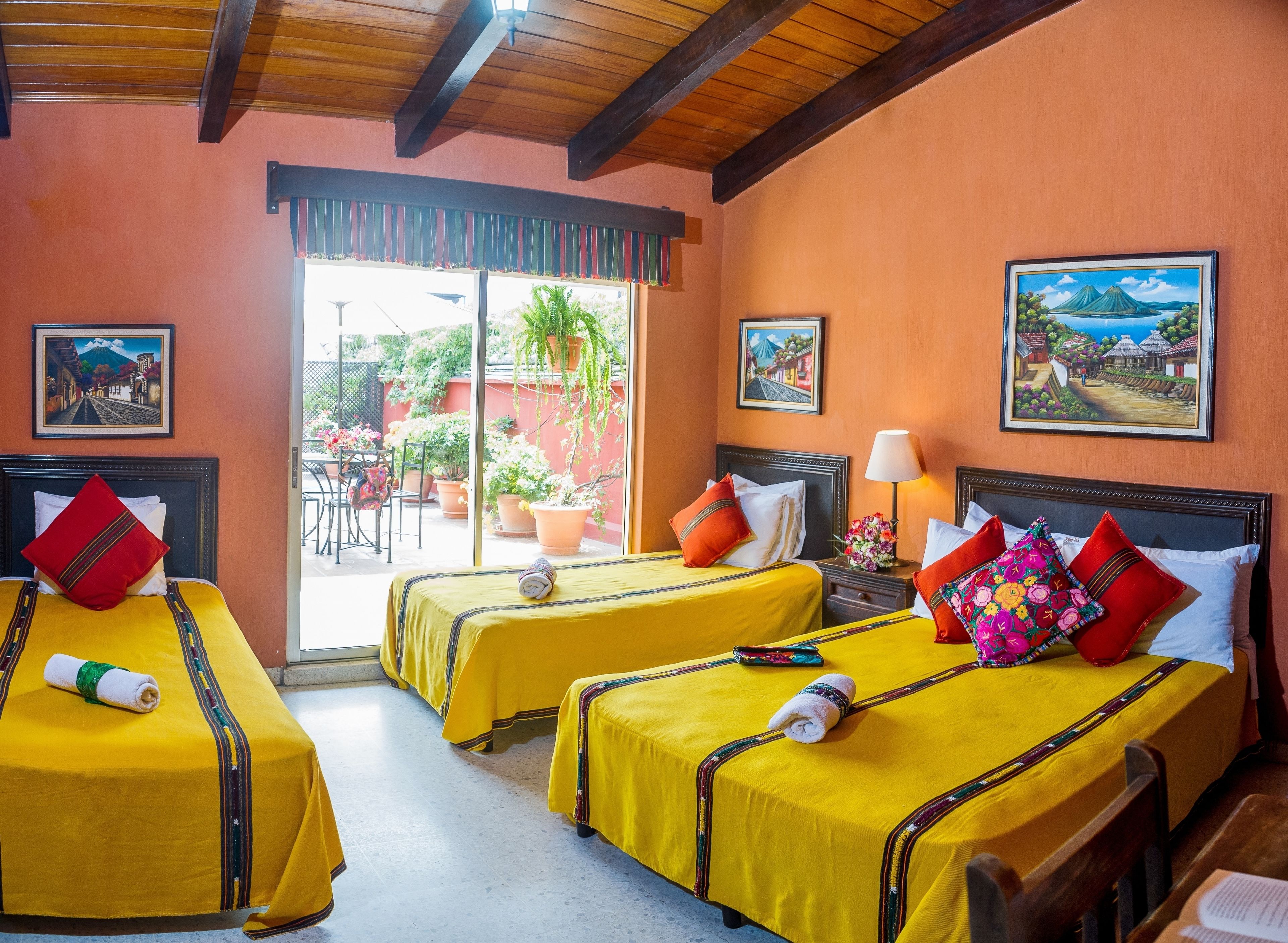 Habitación de hotel con tres camas, cada una con cubrecama y almohadas amarillas, techo de madera y vistas al patio exterior con plantas y una mesa.