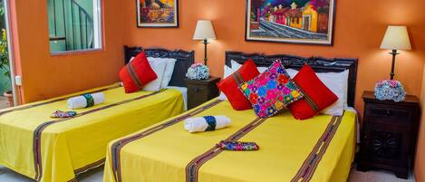 A hotel room with two beds, each with yellow bedding and red pillows, a wooden headboard, and a painting of a colorful street scene on the wall.