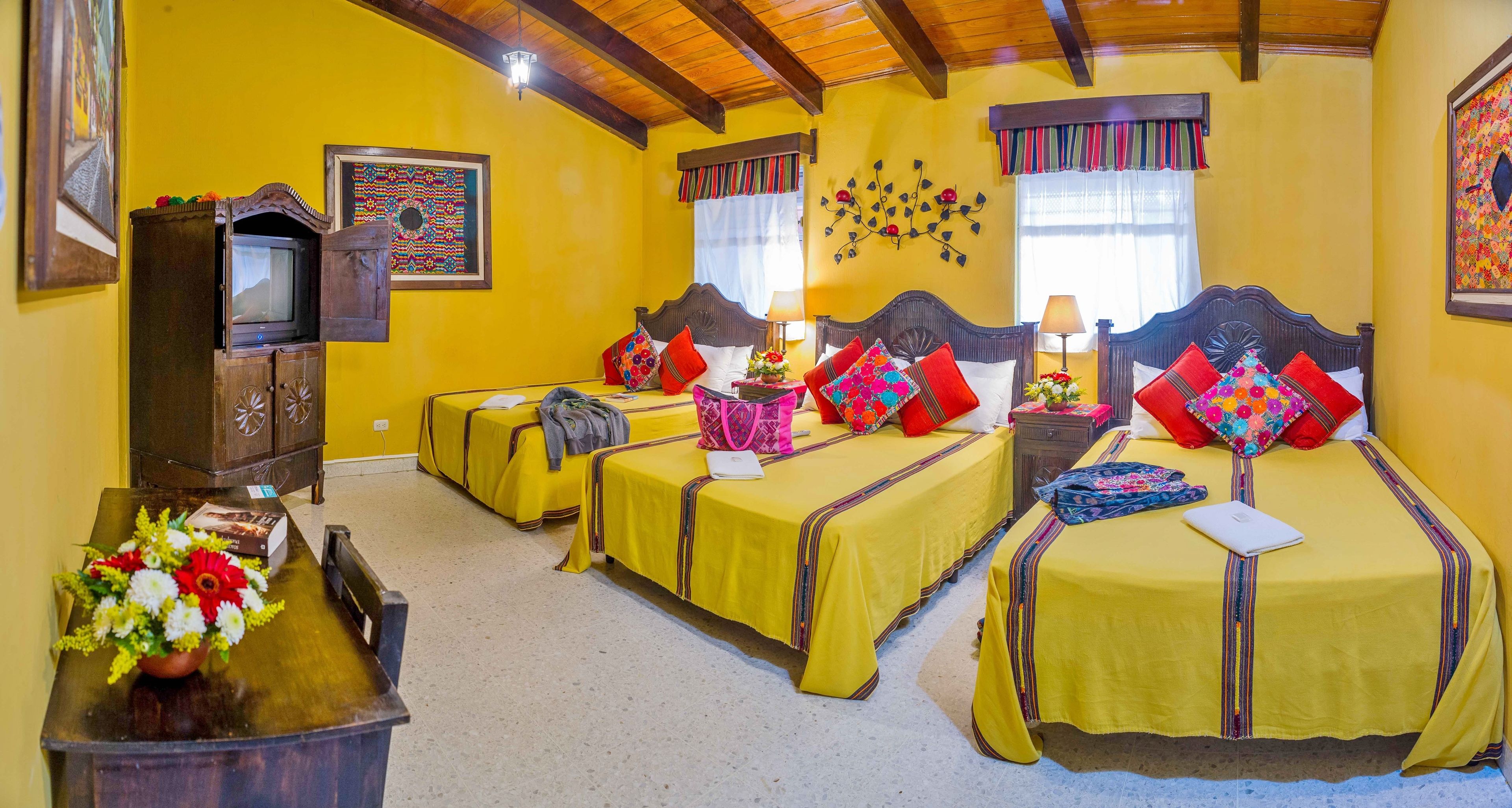 Habitación de hotel con cuatro camas individuales, cada una con cubrecama amarillo y almohadas de colores. Se observa un mueble de madera con un televisor, una mesita con un jarrón de flores y una ventana con cortina decorativa.