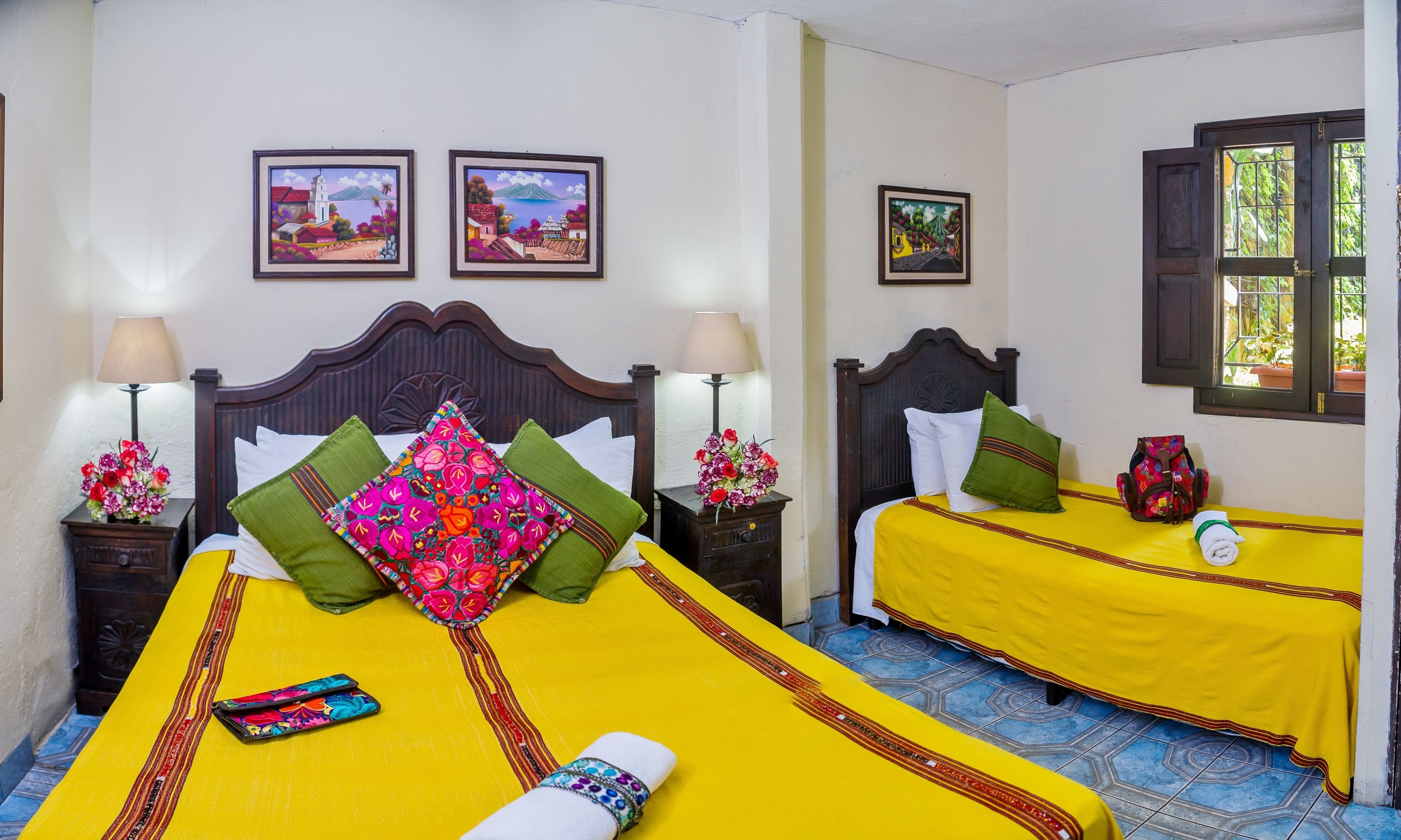 A hotel room with two beds, each with a yellow bedspread and pillows, a wooden headboard, and a nightstand with a lamp and flowers.