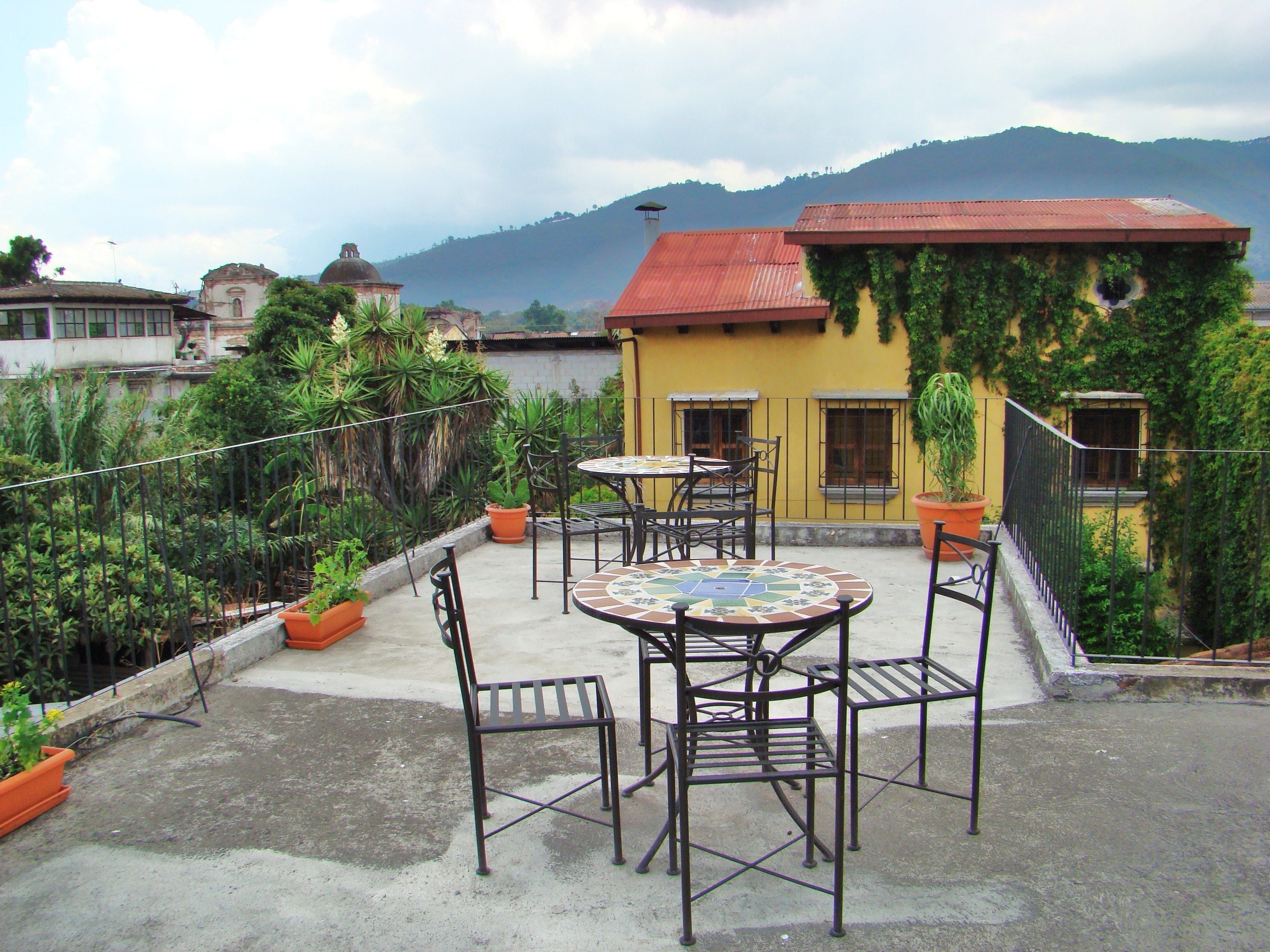 Terraza en la azotea con una mesa redonda y sillas, rodeada de plantas en macetas y vistas a una montaña.