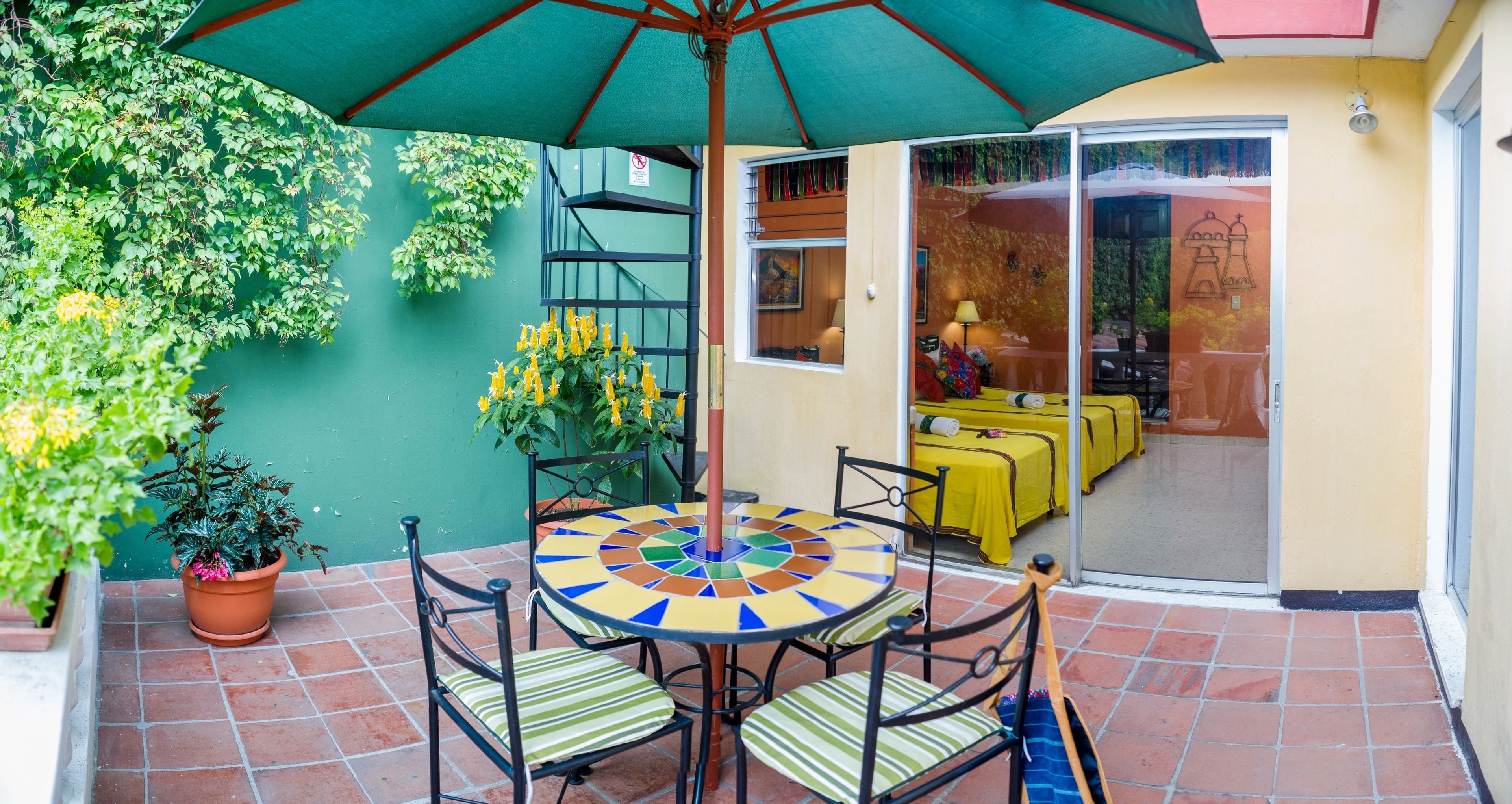 Een terras met een ronde tafel en stoelen, een groene parasol en een gele tafelkleed.