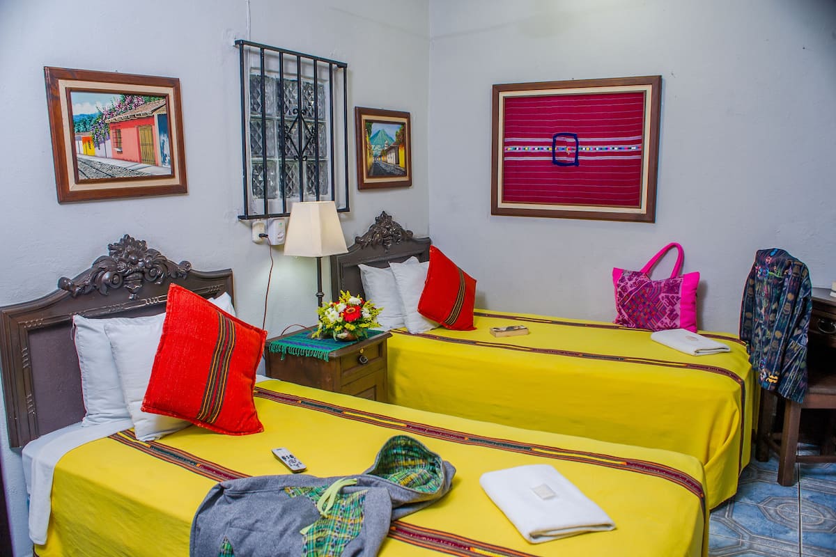 Habitación de hotel con dos camas, cada una con cubrecama amarillo y almohadas rojas. Hay una mesita de noche con una lámpara, un bolso rojo y una chaqueta estampada colgada en la pared.
