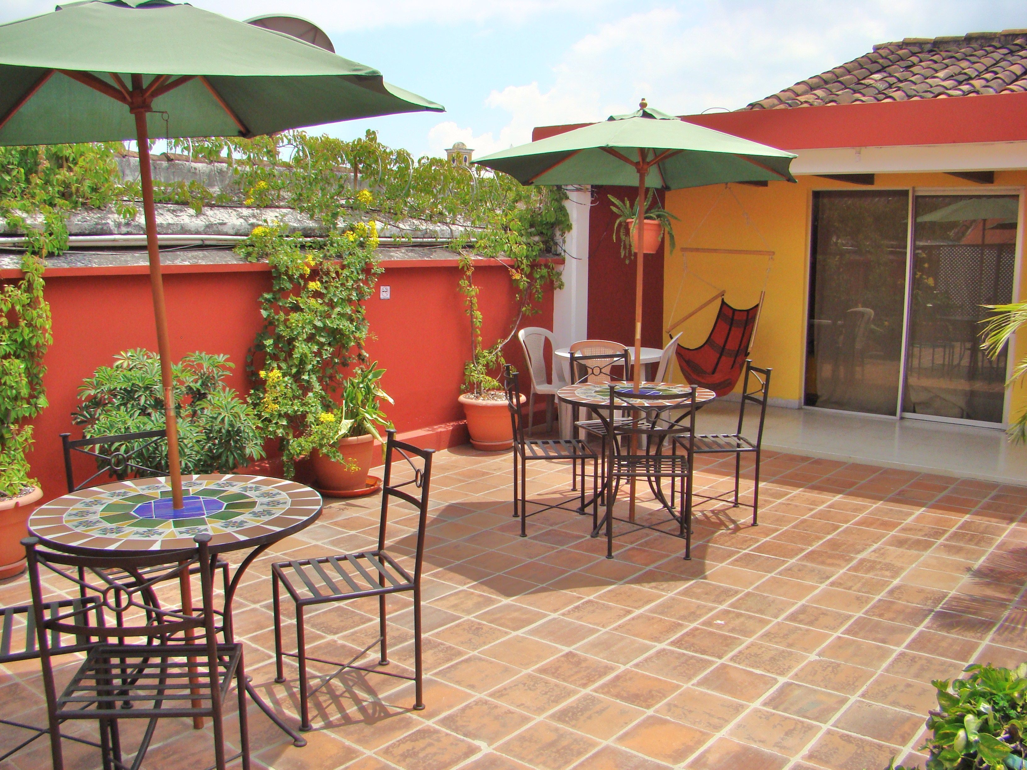 Un patio al aire libre con suelo de baldosas, una mesa redonda con sillas y dos sombrillas que dan sombra.
