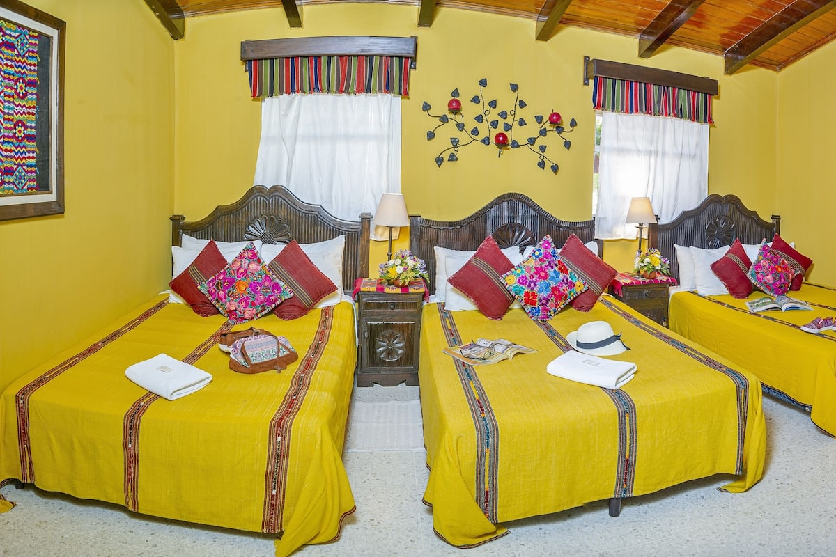 Habitación de hotel con dos camas individuales, cada una con una colcha amarilla y varias almohadas. Se observa un cabecero de madera, una mesita de noche con lámpara, y una pequeña mesa con una cesta. Las paredes son amarillas y el techo es de madera.