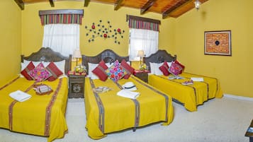 Quarto de hotel com duas camas de solteiro, cada uma com colcha amarela e várias almofadas. Apresenta cabeceira de madeira, mesa de cabeceira com abajur e uma pequena mesa com cesta. O quarto tem paredes amarelas e teto de madeira.