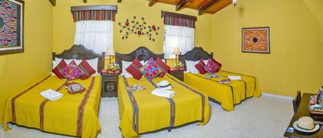 Habitación de hotel con dos camas individuales, cada una con una colcha amarilla y varias almohadas. Se observa un cabecero de madera, una mesita de noche con lámpara, y una pequeña mesa con una cesta. Las paredes son amarillas y el techo es de madera.