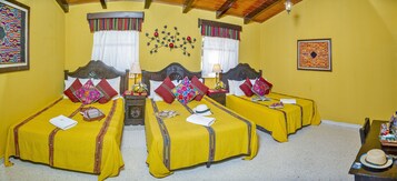 Quarto de hotel com duas camas de solteiro, cada uma com colcha amarela e várias almofadas. Apresenta cabeceira de madeira, mesa de cabeceira com abajur e uma pequena mesa com cesta. O quarto tem paredes amarelas e teto de madeira.