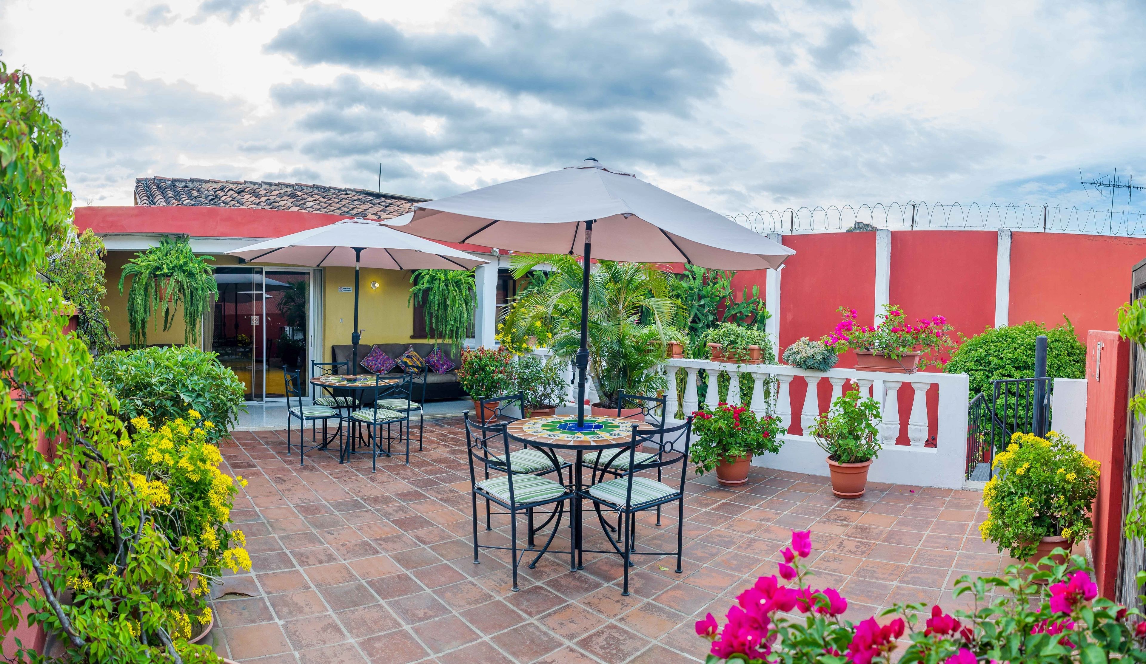 Un patio con una mesa redonda y sillas, rodeado de plantas en macetas y un muro rojo.