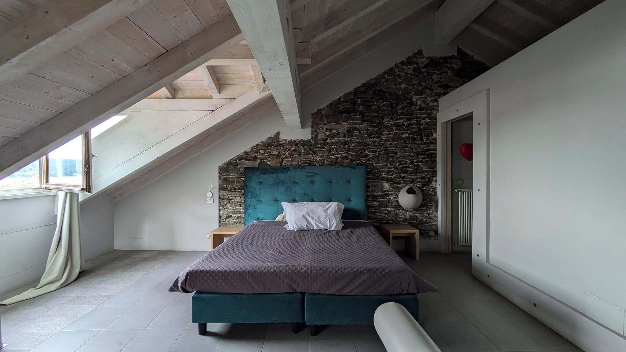 Ein Schlafzimmer mit schrägverdeckter Decke, einer Steinwand, einem Bett mit smaragdgrünem Kopfteil und einem Fenster.