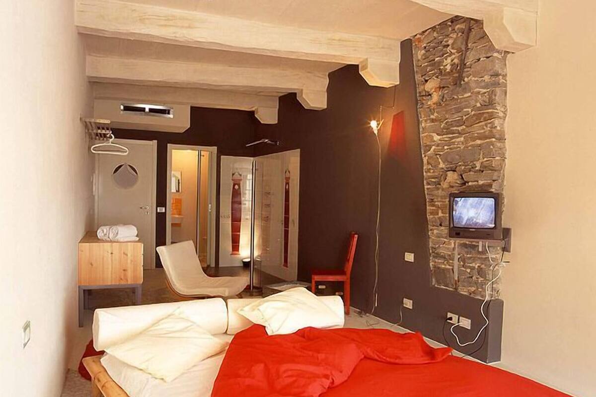 A bedroom with a bed, a red blanket, a stone wall, a television, and a wooden bench.