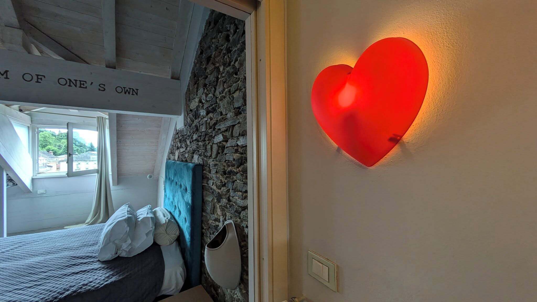 Une chambre avec un mur en pierre, un lit doté d’une tête de lit bleue et un luminaire rouge en forme de cœur fixé au mur.