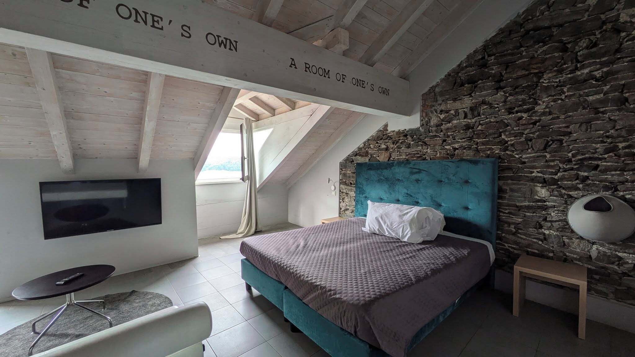 Ein Schlafzimmer mit Stehwänden, einem Bett mit smaragdgrünem Kopfteil, einem an der Wand montierten Fernseher und einem Schild an der Decke mit der Aufschrift 'A ROOM OF ONE'S OWN'.