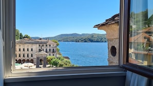 A room with a large window overlooking a lake, a stone bridge, and a building with arched windows. - Hotel Aracoeli (Orta San Giulio)
