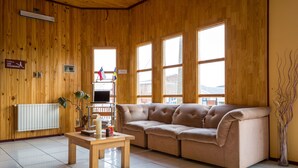 A wooden-paneled room with a beige sectional sofa, a wooden table, and a radiator. - Hostal Alcazar  (Natales)